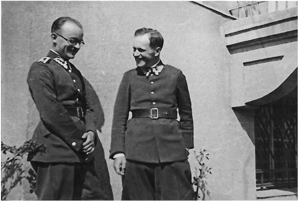 Zdjęcie czarno-białe, dwóch mężczyzn w mudnurach Wojska Polskiego rozmawia na tle ściany budynku.