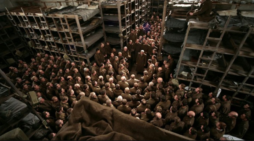 Kadr z filmu Katyń Andrzeja Wajdy z 2007 roku. Duża grupa uwięzionych żołnierzy polskich stoi stłoczona w obozowym baraku. Żołnierze ubrani są w płaszcze oficerskie. Za nimi widać wysokie prycze.