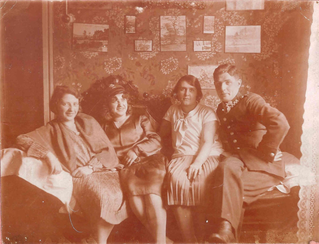 Zdjęcie w sepi, trzy siedzące kobiety i jeden mężczyzna w mundurze, w tle obrazki na ścianie.