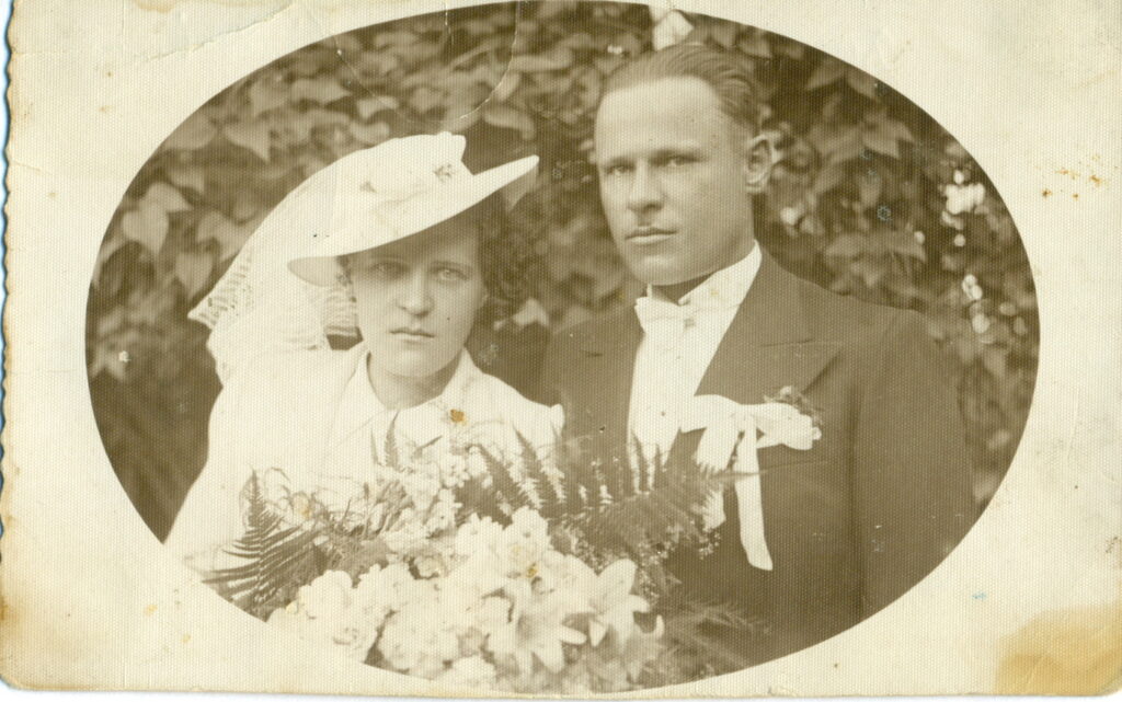 Zdjęcie czarno-białe, ślubne. Kobieta w białym kapeluszu i z bukietem kwiatów, mężczyzna w garniturze.